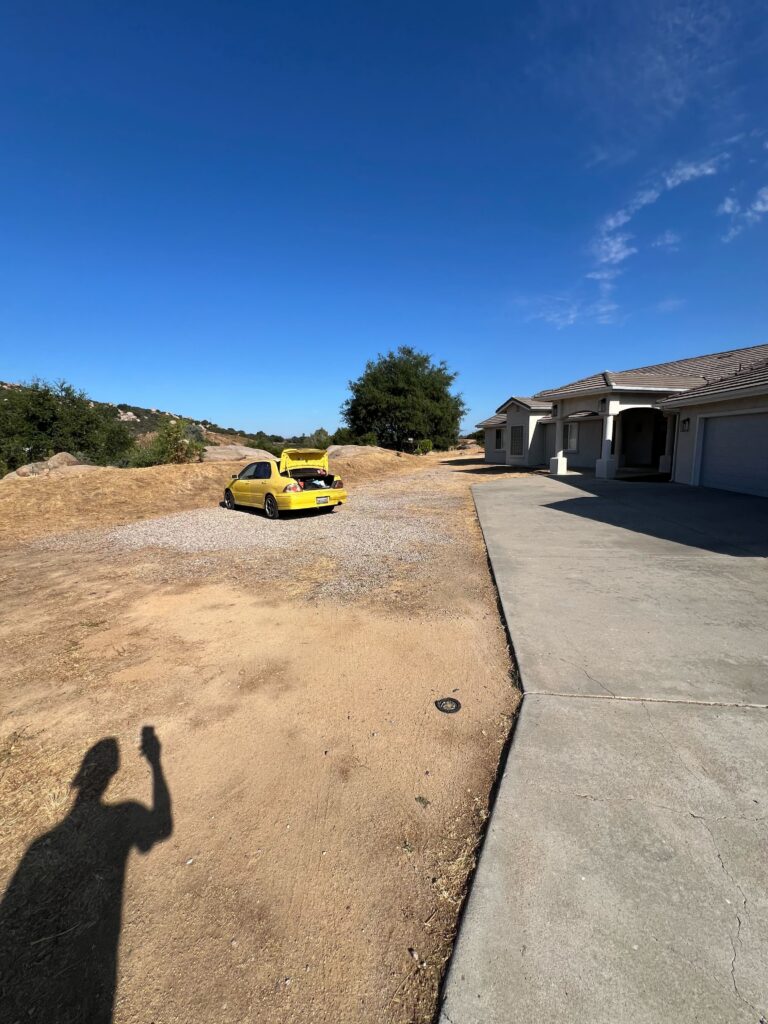 Front yard of a residential property showing a large expanse of bare, compacted soil adjacent to an existing concrete...