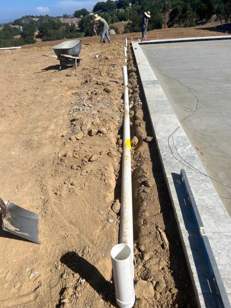 PVC perimeter drain pipe laid in an open trench running parallel to a poured concrete pool deck. Workers with shovels and...