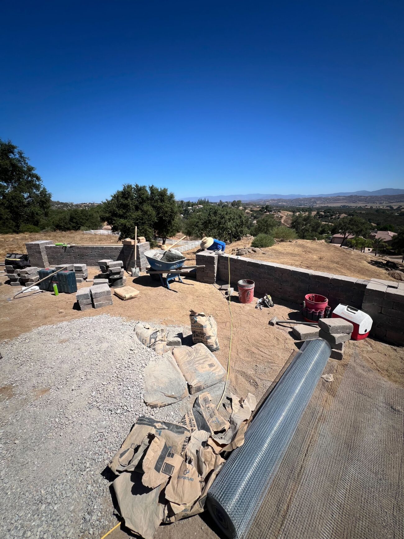 Backyard site with materials staged for a paver patio: stacks of interlocking pavers and a sand/gravel base alongside a...