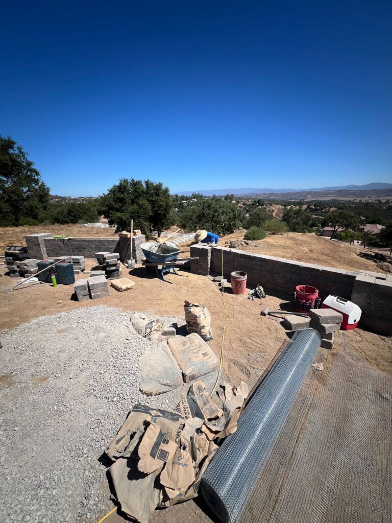 Backyard site with materials staged for a paver patio: stacks of interlocking pavers and a sand/gravel base alongside a...