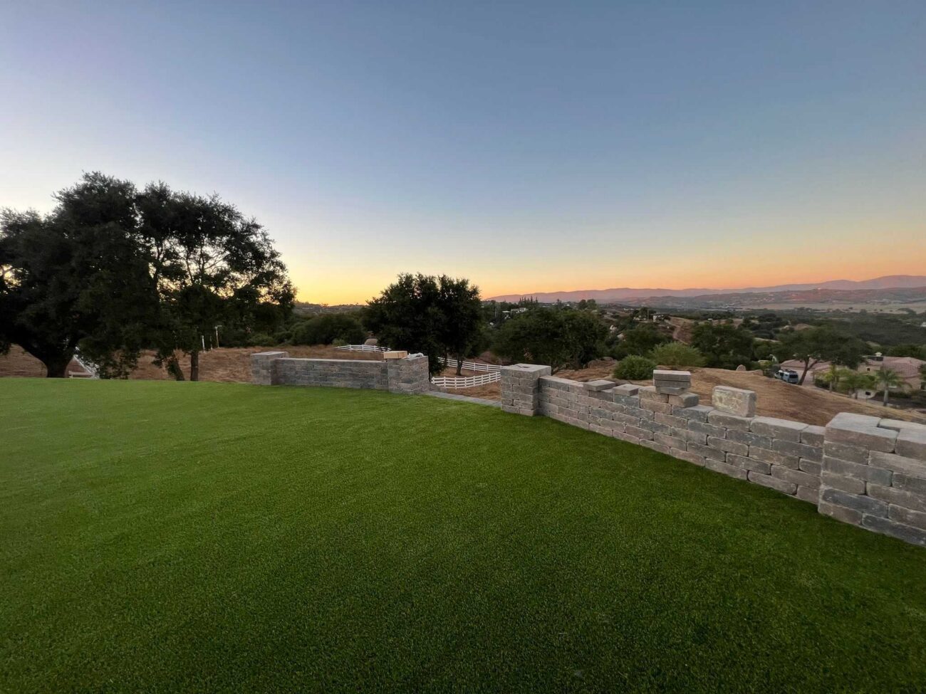Completed residential backyard showing newly installed artificial turf and a segmental retaining wall with stone...