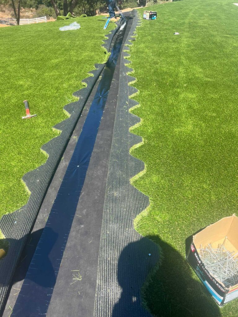 Artificial turf installation in progress showing a long seam with seam tape/underlayment exposed and turf edges trimmed...