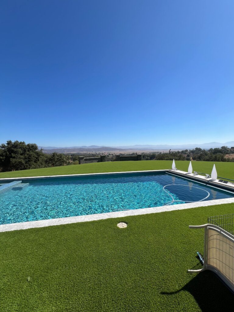 Residential backyard showing a completed artificial turf installation surrounding an in-ground pool with visible stone...