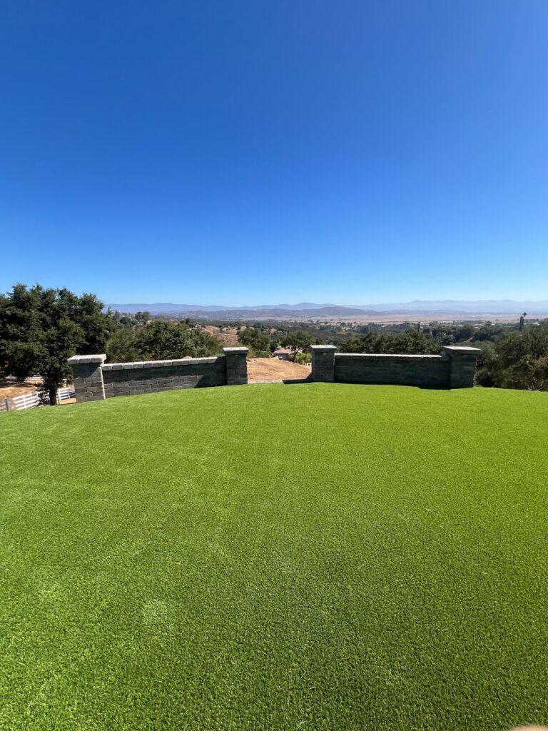 Finished artificial turf installed across a residential backyard with a low segmental retaining wall and capped pillars...