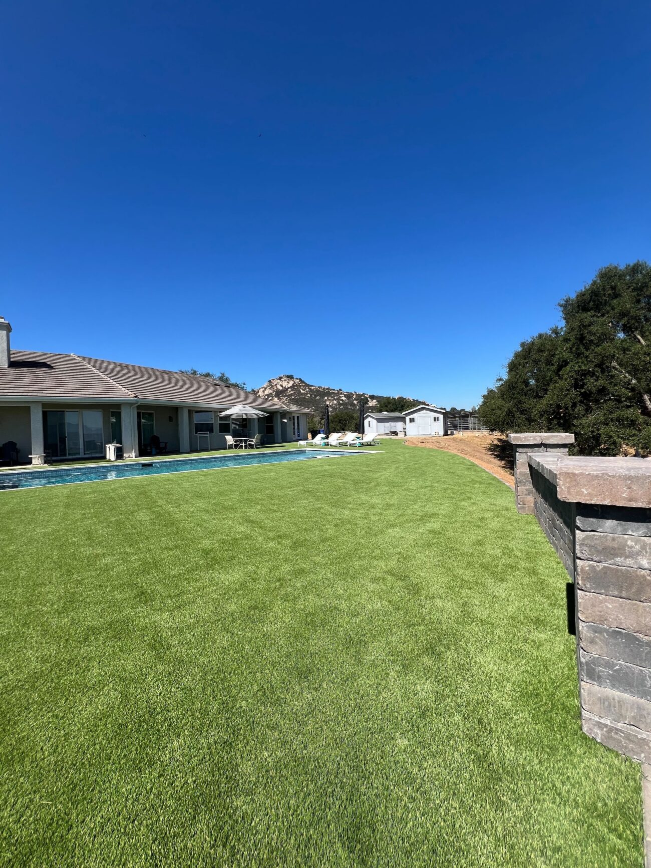 Finished artificial turf lawn installed adjacent to a residential pool with stone wall and capped pillars along the...