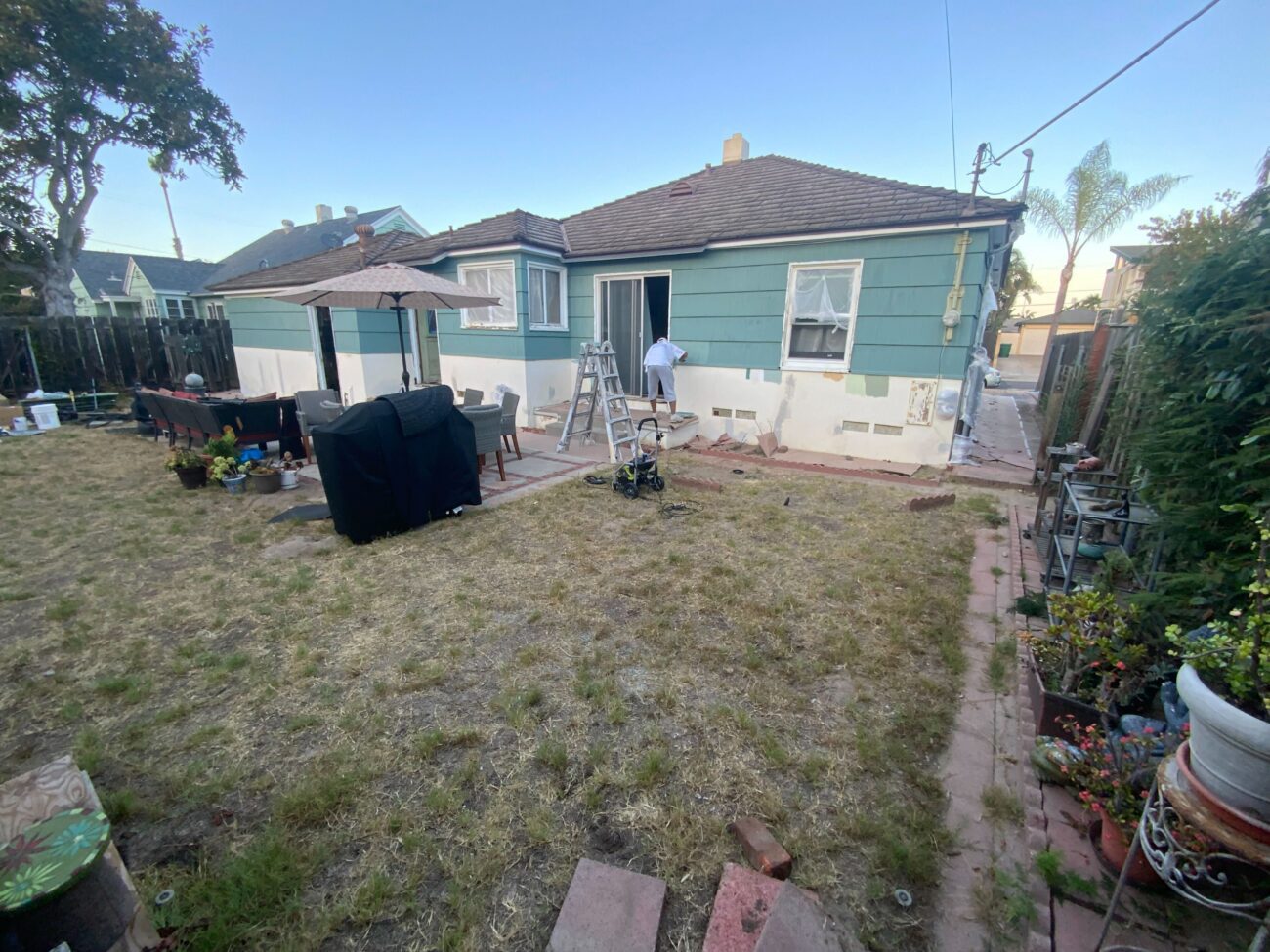 Backyard site photo showing a small paver patio and a paver walkway adjacent to a single-story house, with patchy natural...