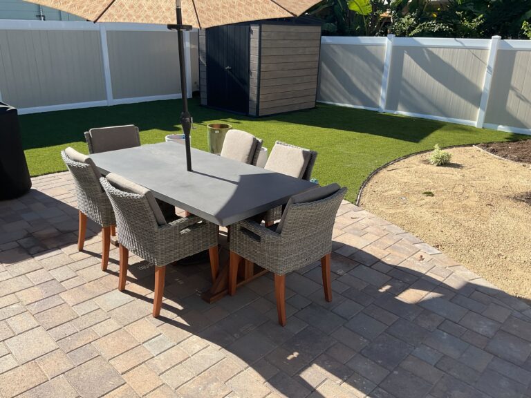 Finished interlocking paver patio with an outdoor dining set and umbrella adjacent to newly installed synthetic turf. A...