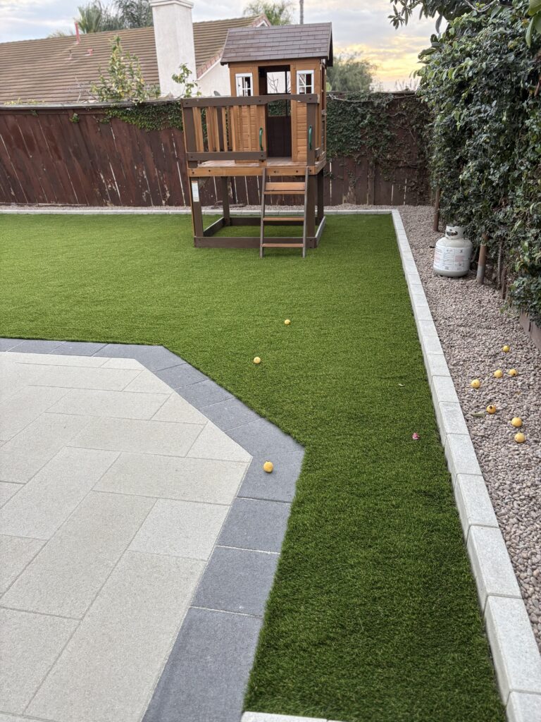 Residential backyard showing newly installed artificial turf abutting an interlocking paver patio with a contrasting dark...