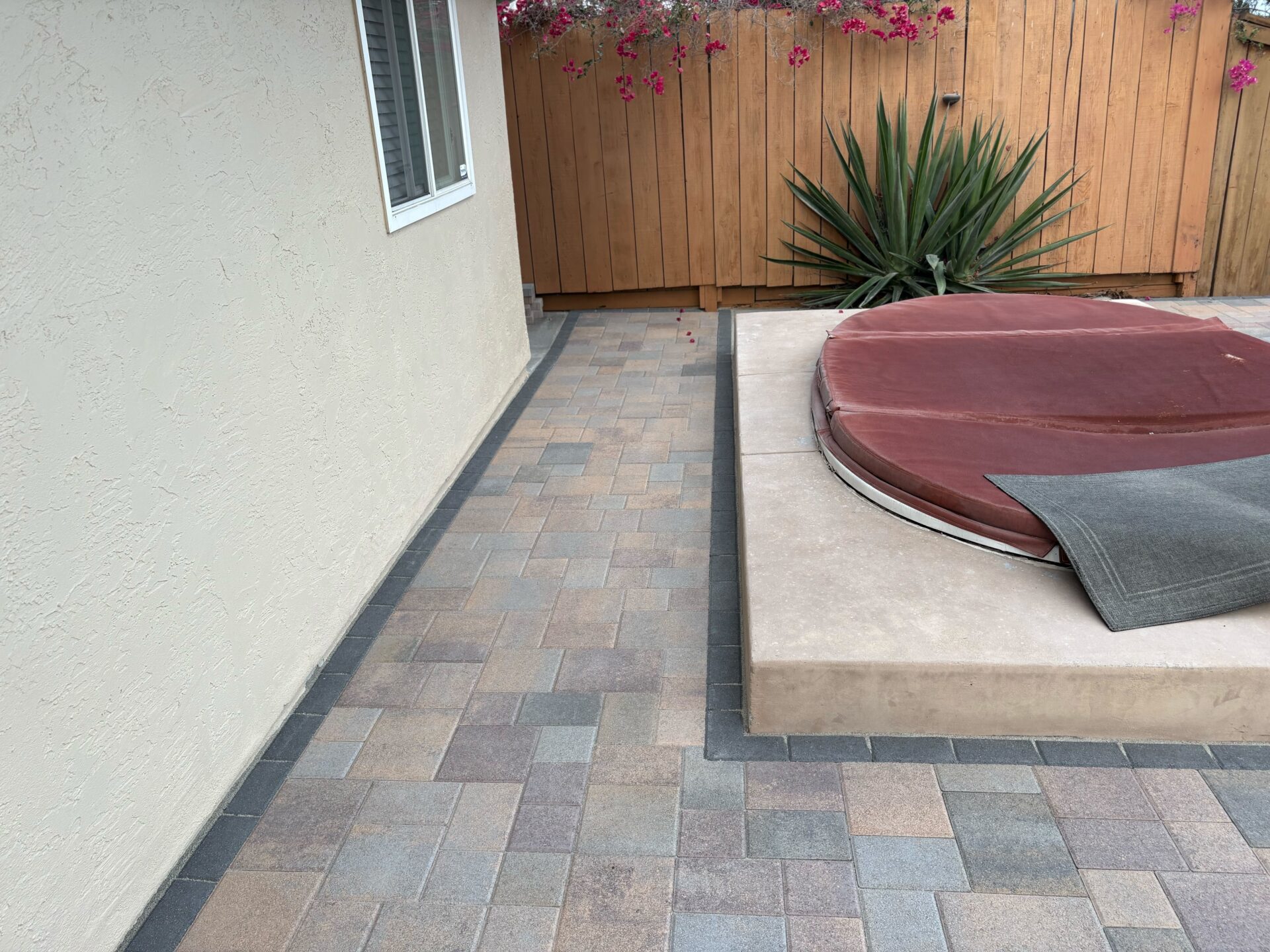 Finished interlocking pavers installed in a side/back yard creating a walkway and patio area around a raised spa. Dark...