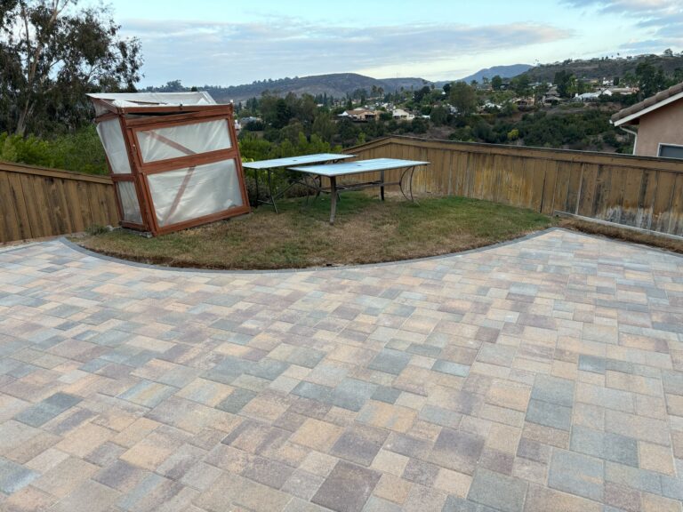 Large interlocking concrete paver patio in multi-tone finish covers most of a residential backyard. A small pocket of...