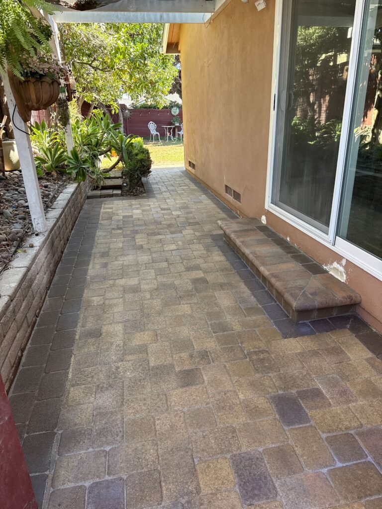 Interlocking concrete pavers form a finished patio/walkway running along the exterior of a residence, adjacent to a...