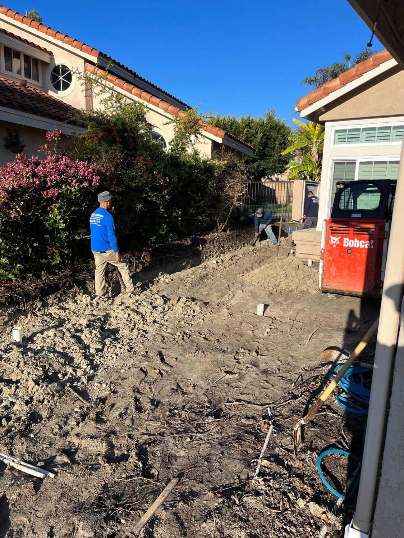 Crew + Bobcat in graded backyard with plumbing risers. Documents the backyard demo phase with people/equipment scale and...