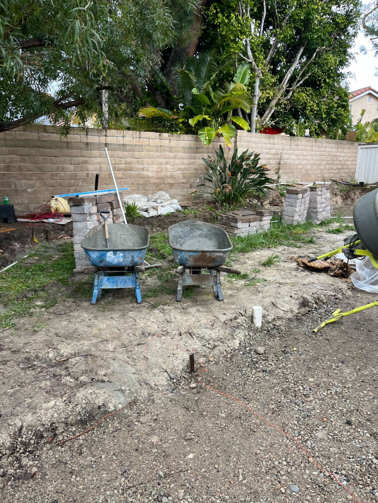 Mortar mix wheelbarrows staged with masonry block stacked at the back wall — proves the wall-build phase distinct from...