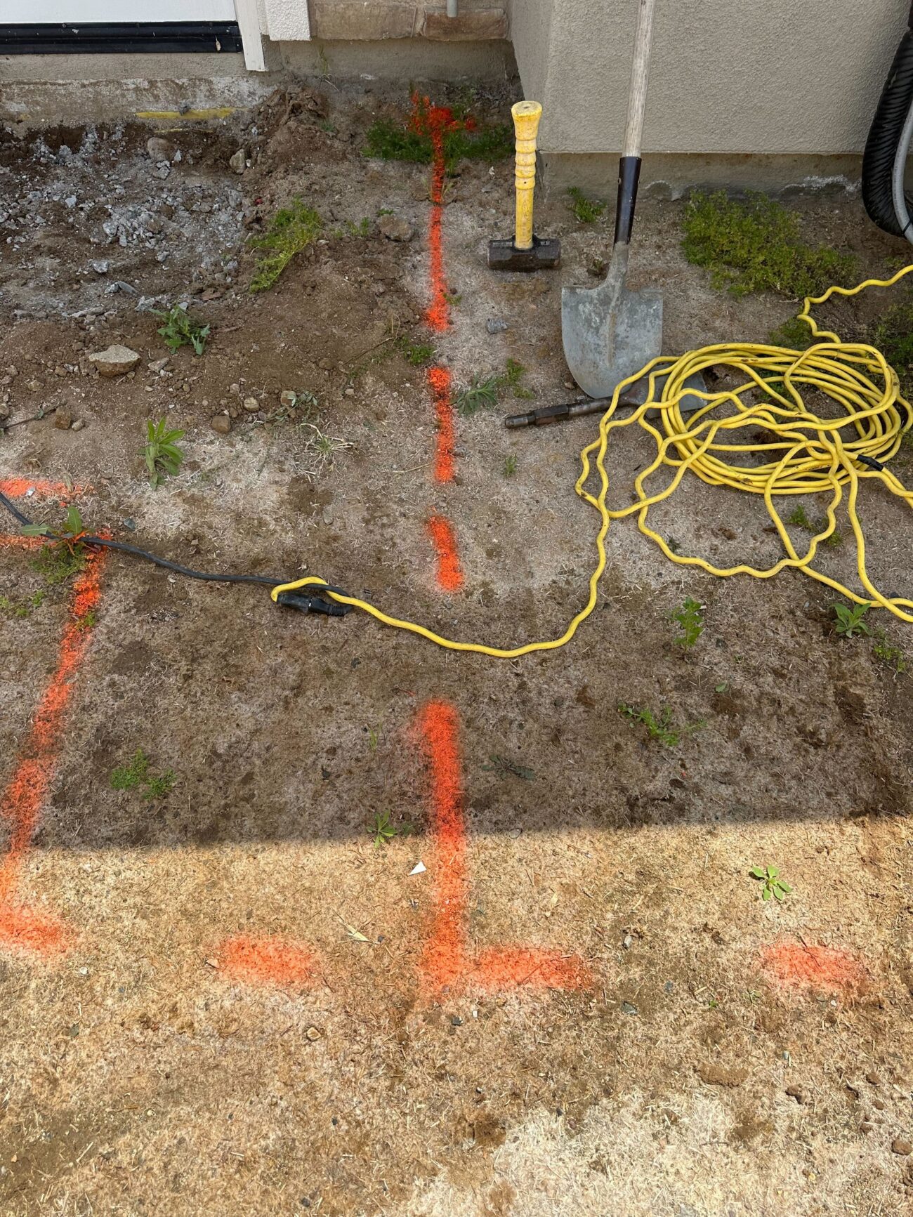 Overview of a residential yard showing orange spray-painted layout lines and hand tools at the house exterior. Exposed...