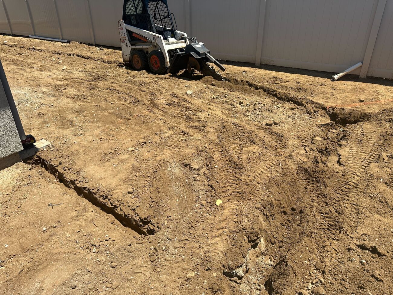 Backyard stripped to subgrade with multiple shallow trenches and track marks visible; a skid-steer loader with a...