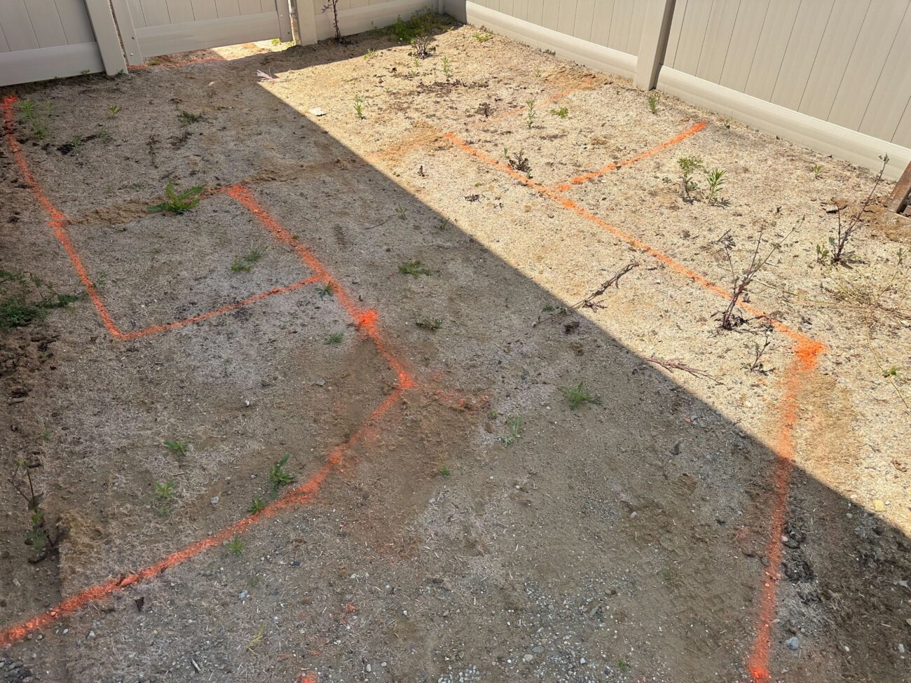 Small fenced backyard showing bare sandy soil with orange spray-painted layout lines marking planned work areas. The...
