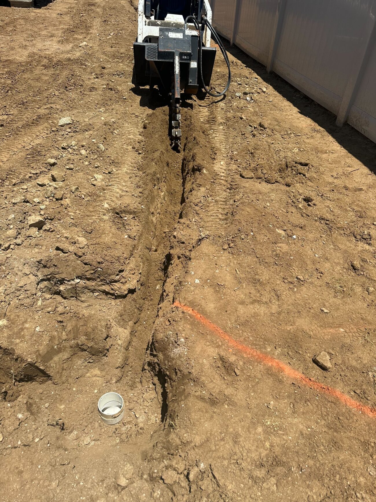 A trencher is cutting a narrow utility/drainage trench through exposed soil in a residential side yard next to a vinyl...