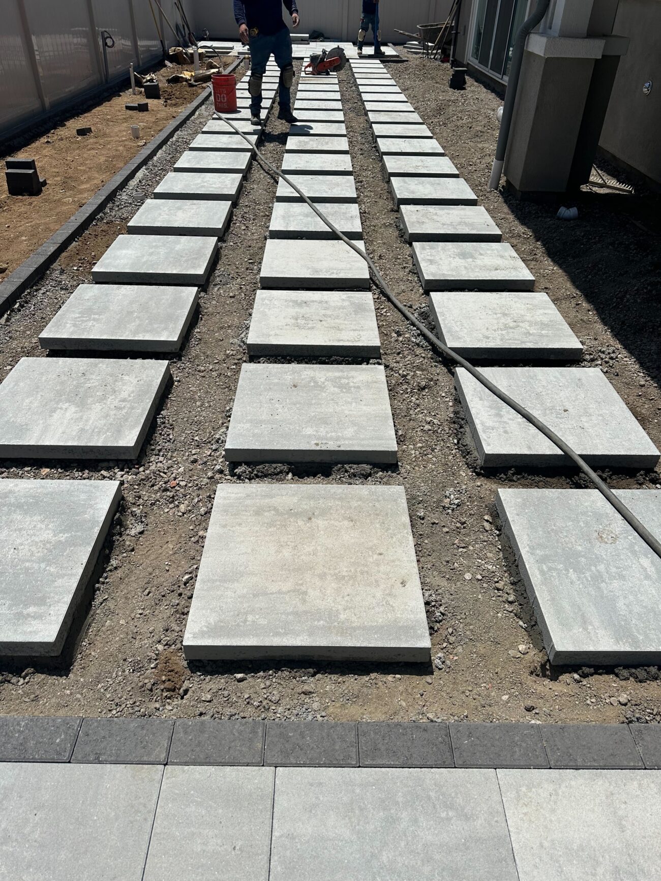 Rows of square concrete pavers are being dry-laid on a compacted aggregate base to form a linear walkway/patio area;...