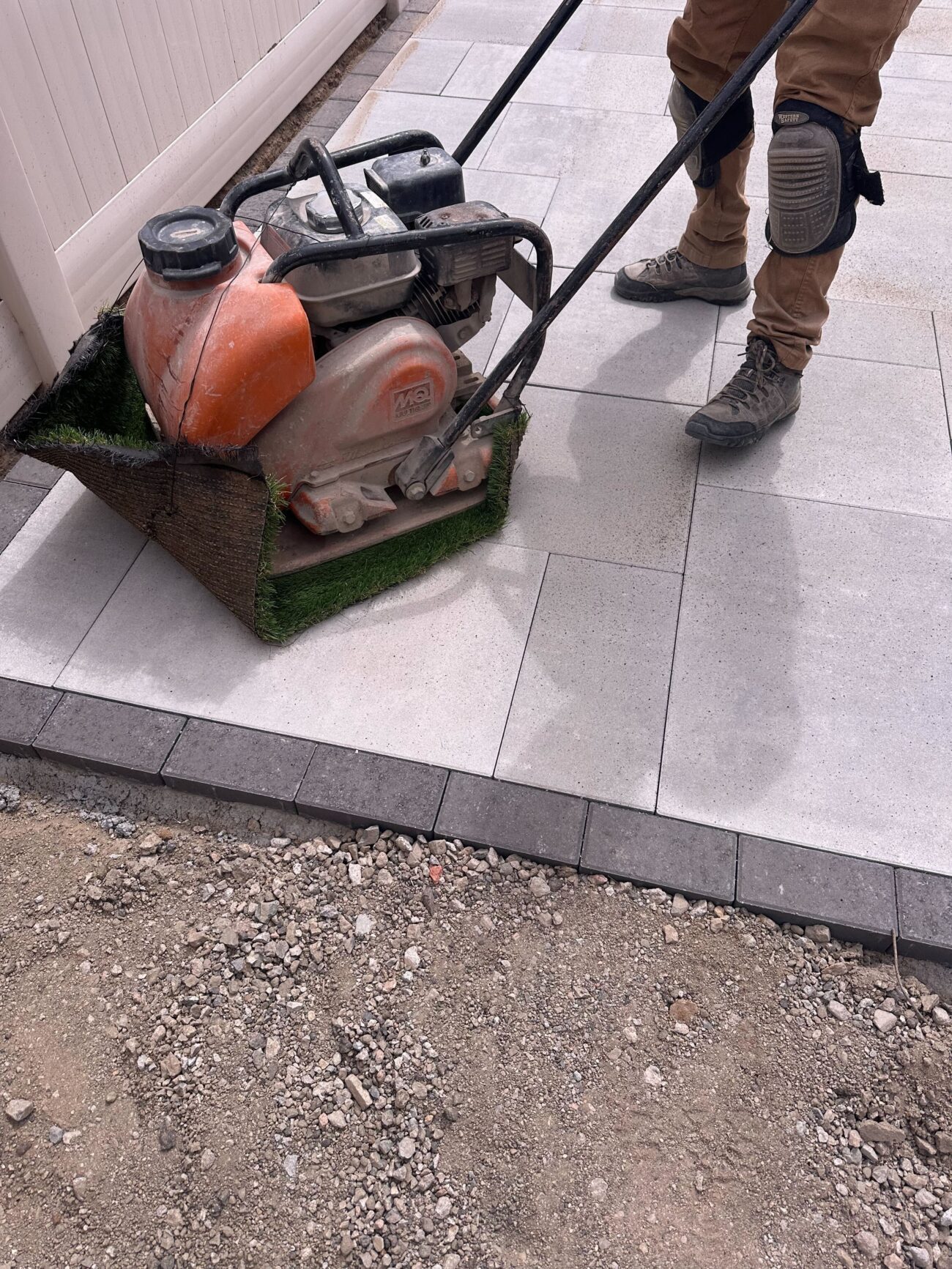 Worker operating a vibratory plate compactor on large-format concrete pavers forming a patio/walkway. An edge restraint...