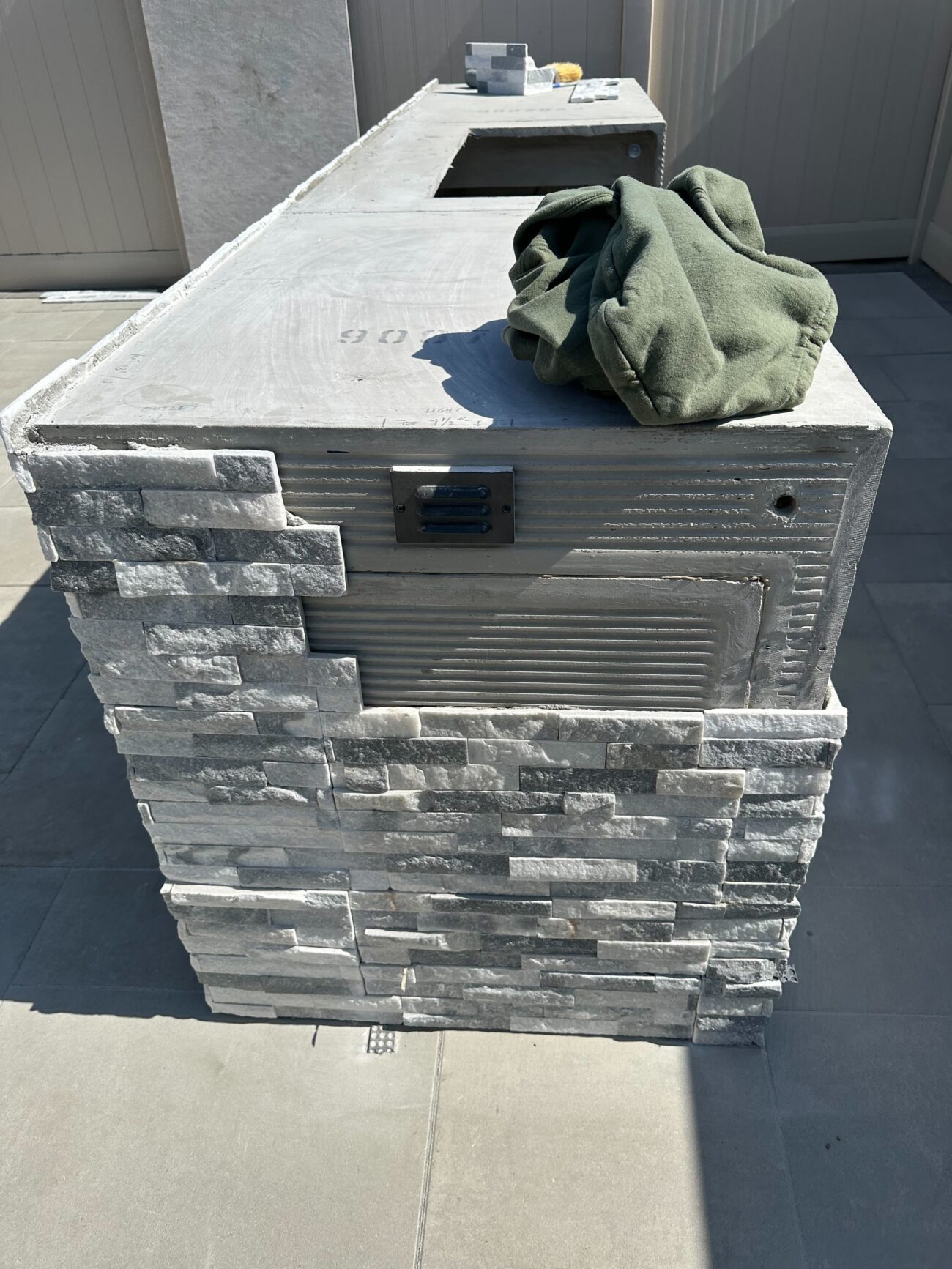 Outdoor kitchen island under active construction with stacked stone veneer partially installed on a concrete base....