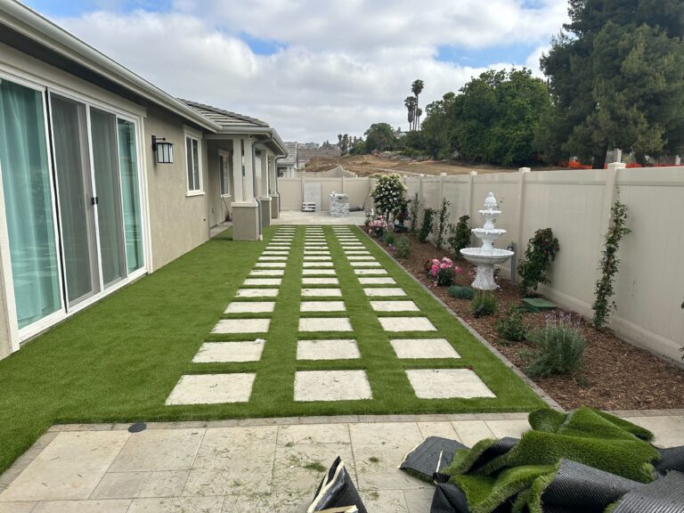 Finished residential side yard showing artificial turf with regularly spaced concrete paver stepping stones forming a...