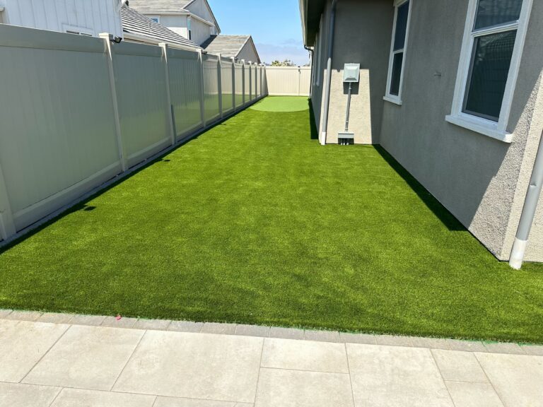 Finished narrow side yard showing newly installed artificial turf bordered by a concrete paver patio/edge and vinyl...