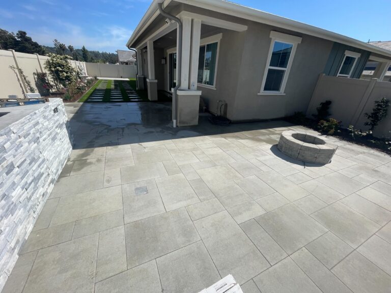Finished outdoor living area showing a large gray interlocking paver patio with a round segmental fire pit and a stacked...