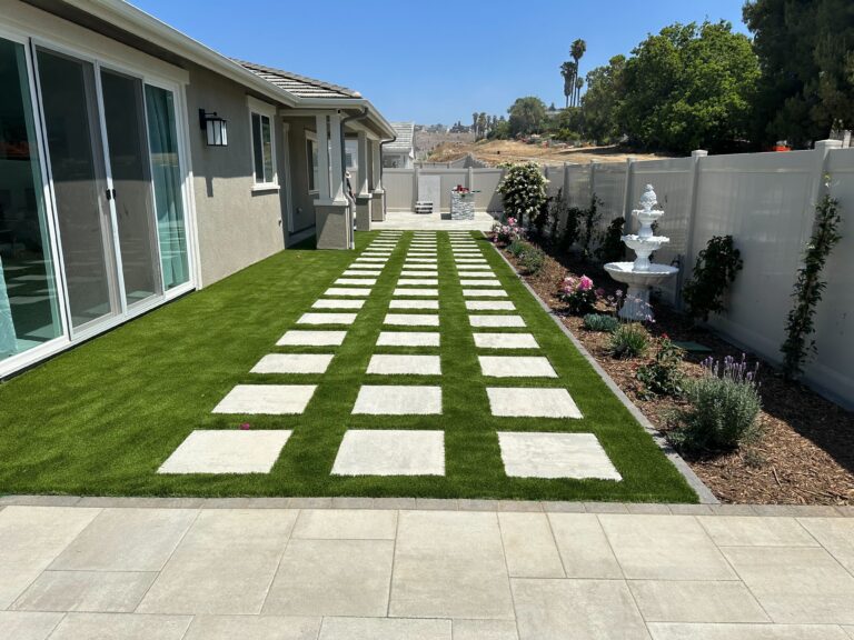 Finished backyard showing a completed landscape with large square paver pads set in artificial turf forming a grid...