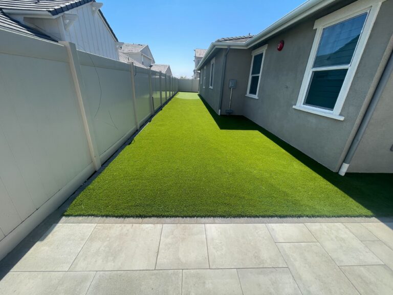 Finished narrow side yard showing newly installed artificial turf running between a vinyl privacy fence and the house,...