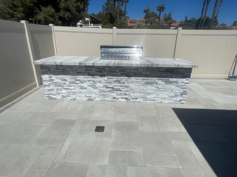 Finished paver patio using large-format light gray pavers with clean joints and a stone-veneer outdoor kitchen island....