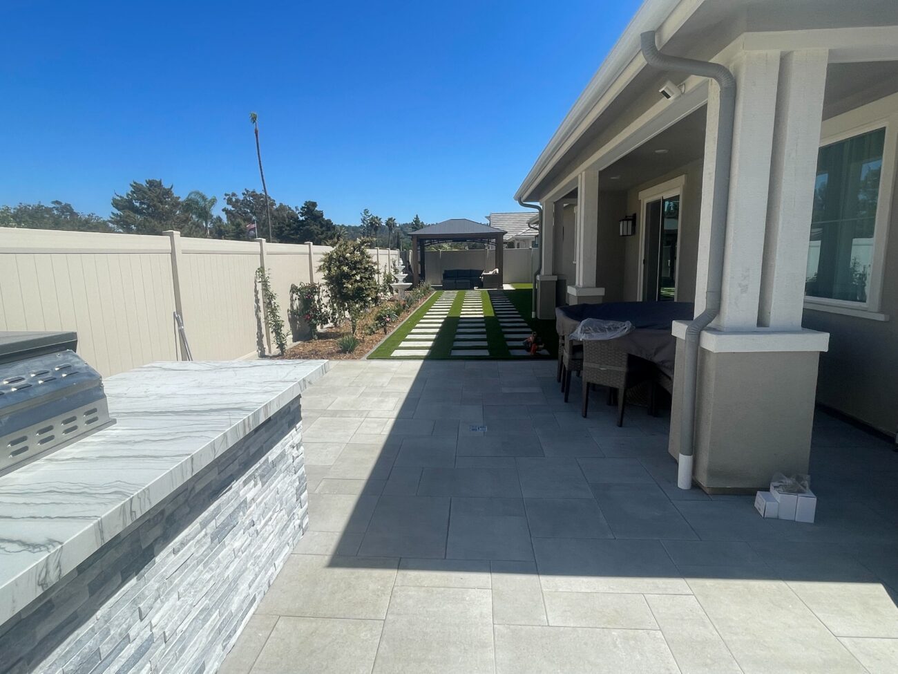 Finished backyard patio showing large light-colored interlocking pavers forming a dining/entertainment area adjacent to a...