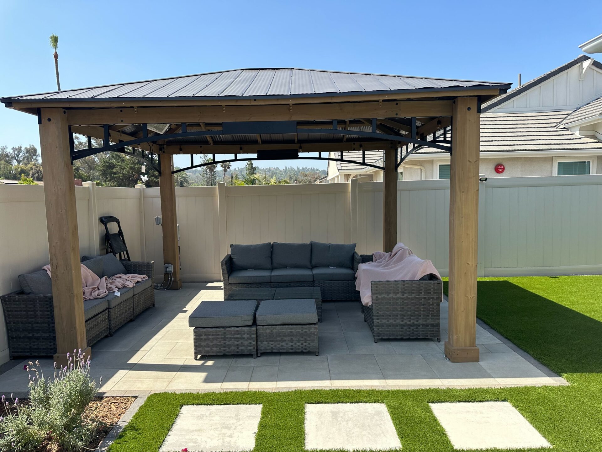 Finished paver patio installed beneath a wood pergola featuring sectional outdoor seating, with adjacent artificial turf...