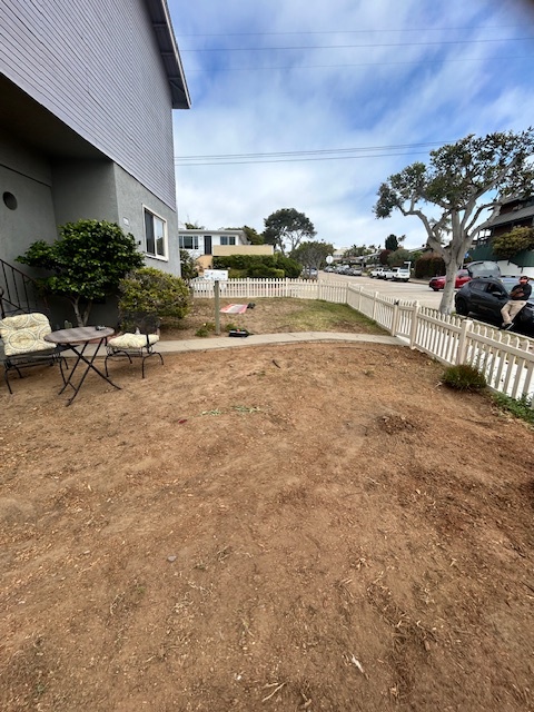 Front yard of a residential property cleared to mostly bare soil with a few shrubs along the house and a white picket...