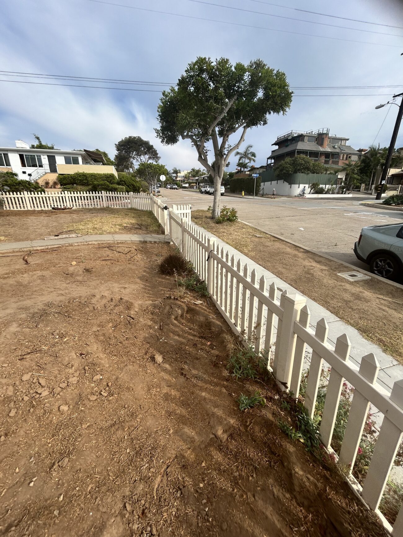 Front yard cleared to bare soil inside a white picket fence with an existing street-facing tree; site appears staged for...