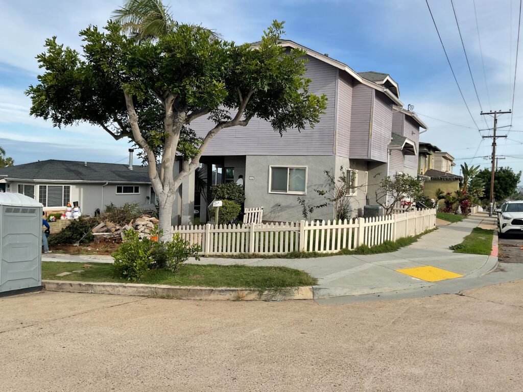 Corner-lot residential front yard undergoing demolition/site prep with disturbed soils, debris piles and a portable...