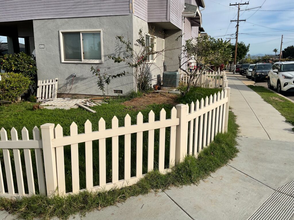 Corner front yard with a white picket fence showing patchy natural turf and exposed soil near the house; an AC condenser...