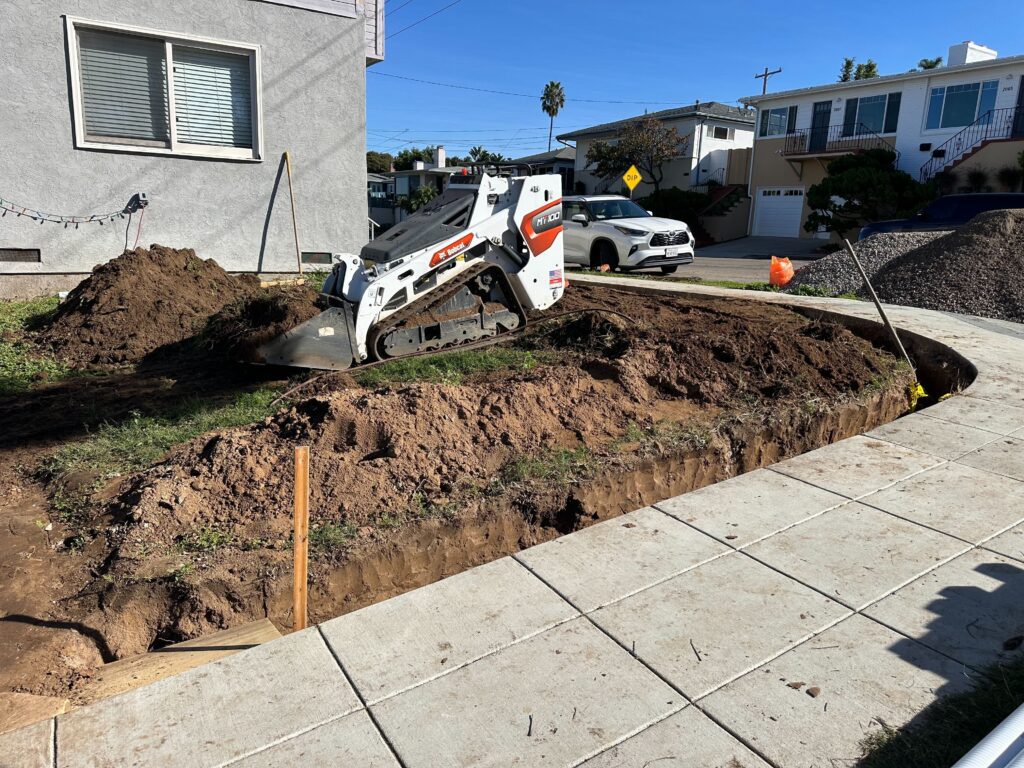 Compact track loader (Bobcat) excavating a residential front yard with trenches and spoil piles adjacent to the concrete...