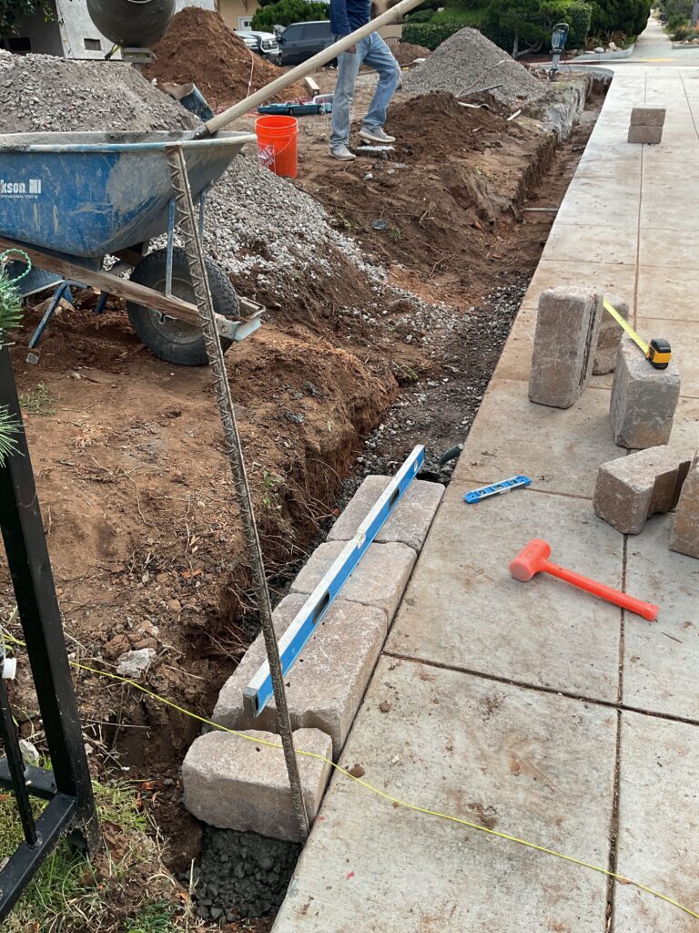 Image shows workers installing a segmental retaining wall along a concrete sidewalk with a trench excavated and base...