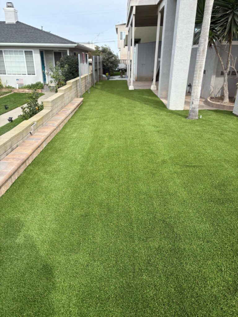 Installed artificial turf covering a narrow side yard adjacent to a residential exterior, flanked by a segmental...