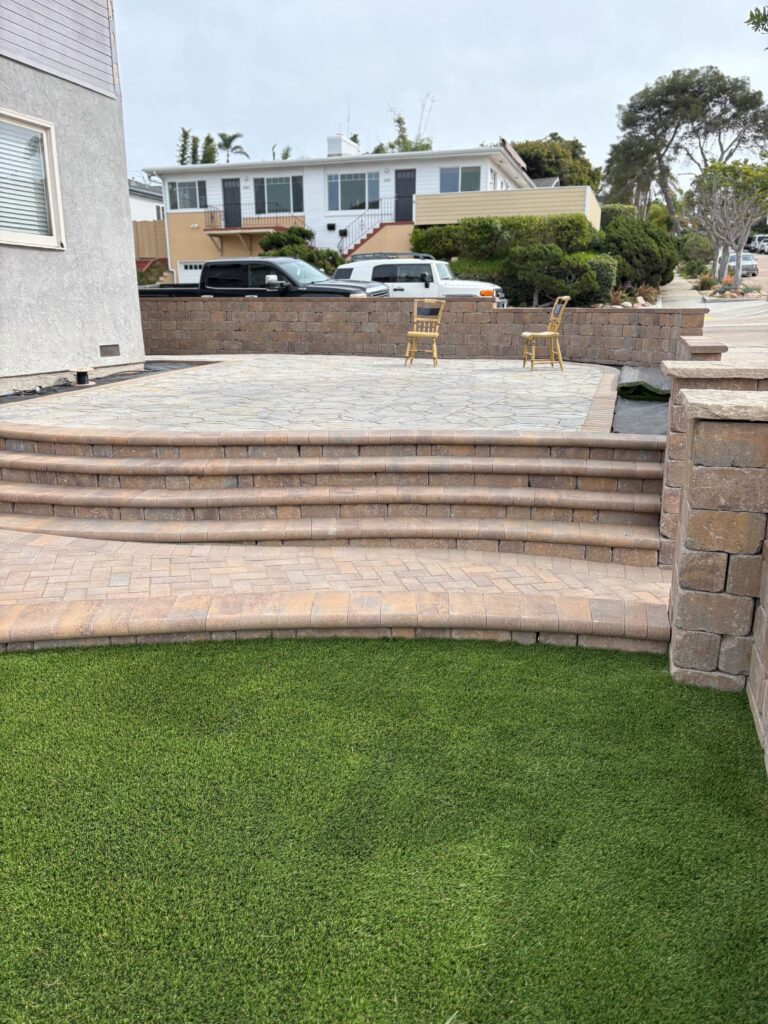 Completed paver patio with multiple riser steps and segmental retaining walls overlooking a synthetic turf lawn....