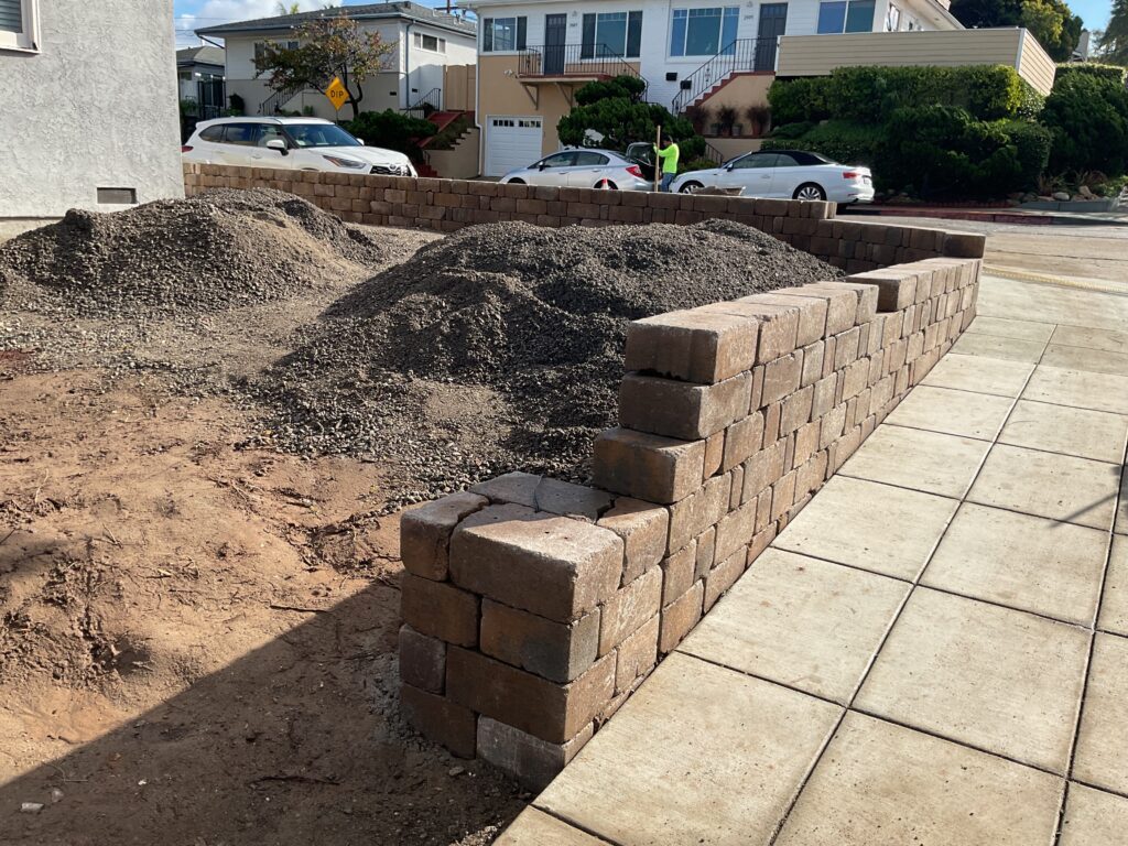 New segmental retaining wall constructed from modular concrete blocks runs along the sidewalk with multiple piles of...