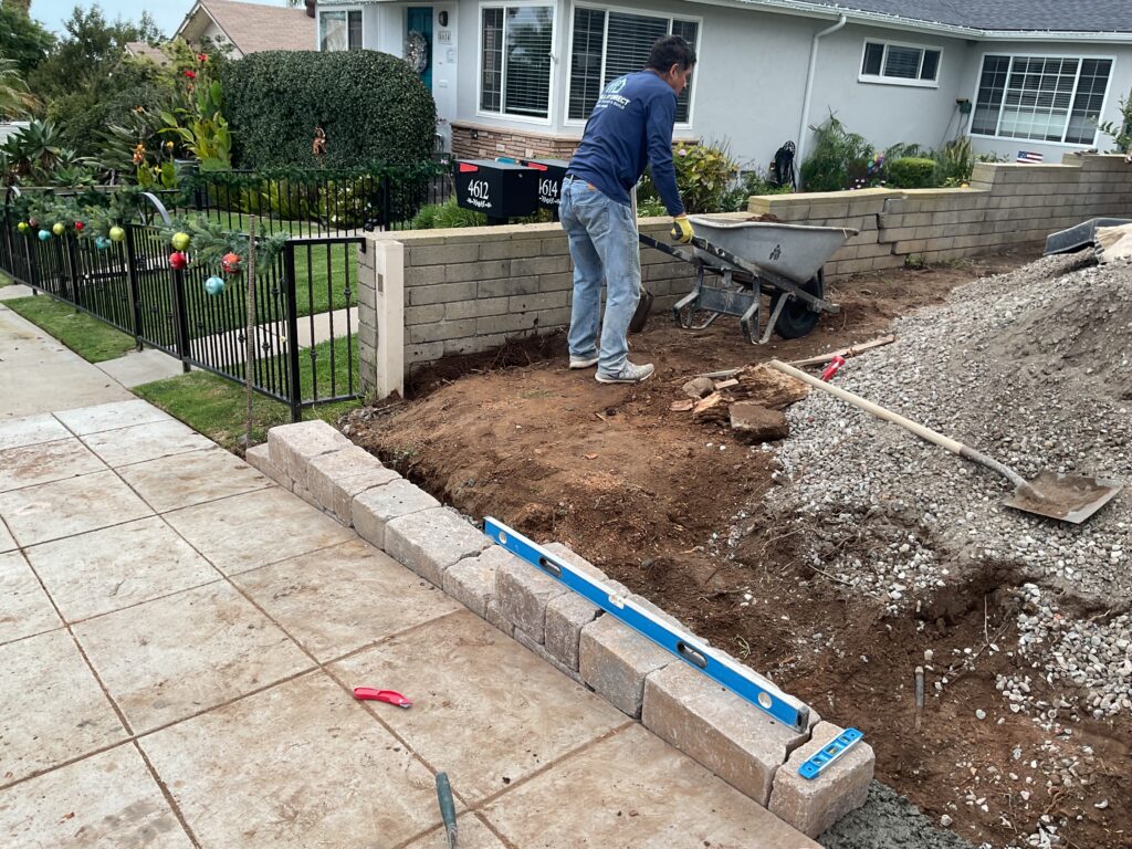 Front-yard construction showing a crew member using a wheelbarrow while preparing a single-course segmental retaining...