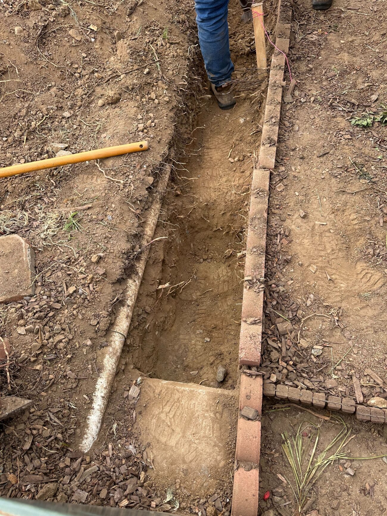 Excavated trench runs parallel to a removed interlocking paver edge in a residential yard. A crew member stands in the...