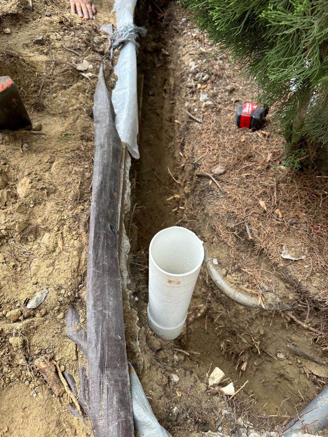 Open trench excavated alongside a planting bed showing a perforated drain pipe wrapped in filter fabric and a vertical...