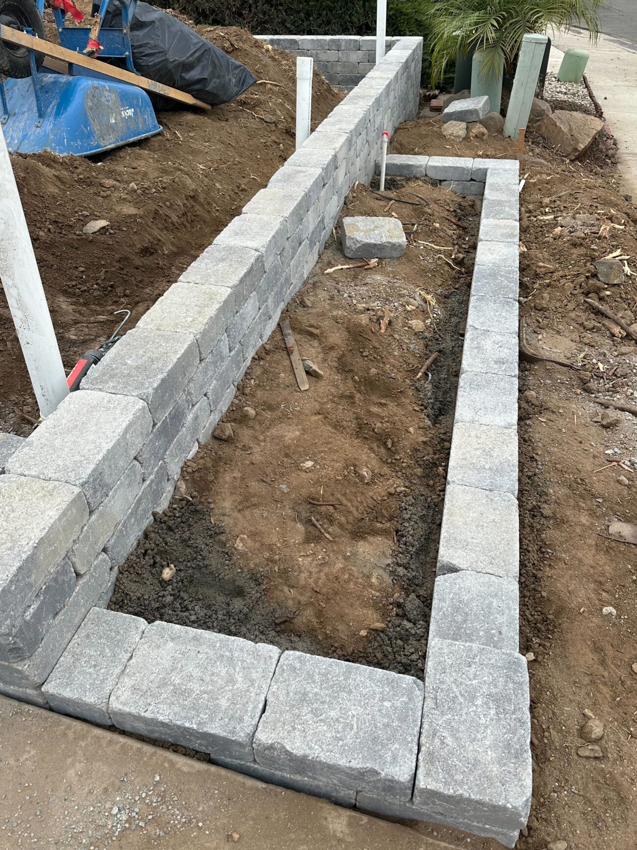 Segmental concrete block retaining wall under active installation forming a narrow planting bed; multiple courses of...