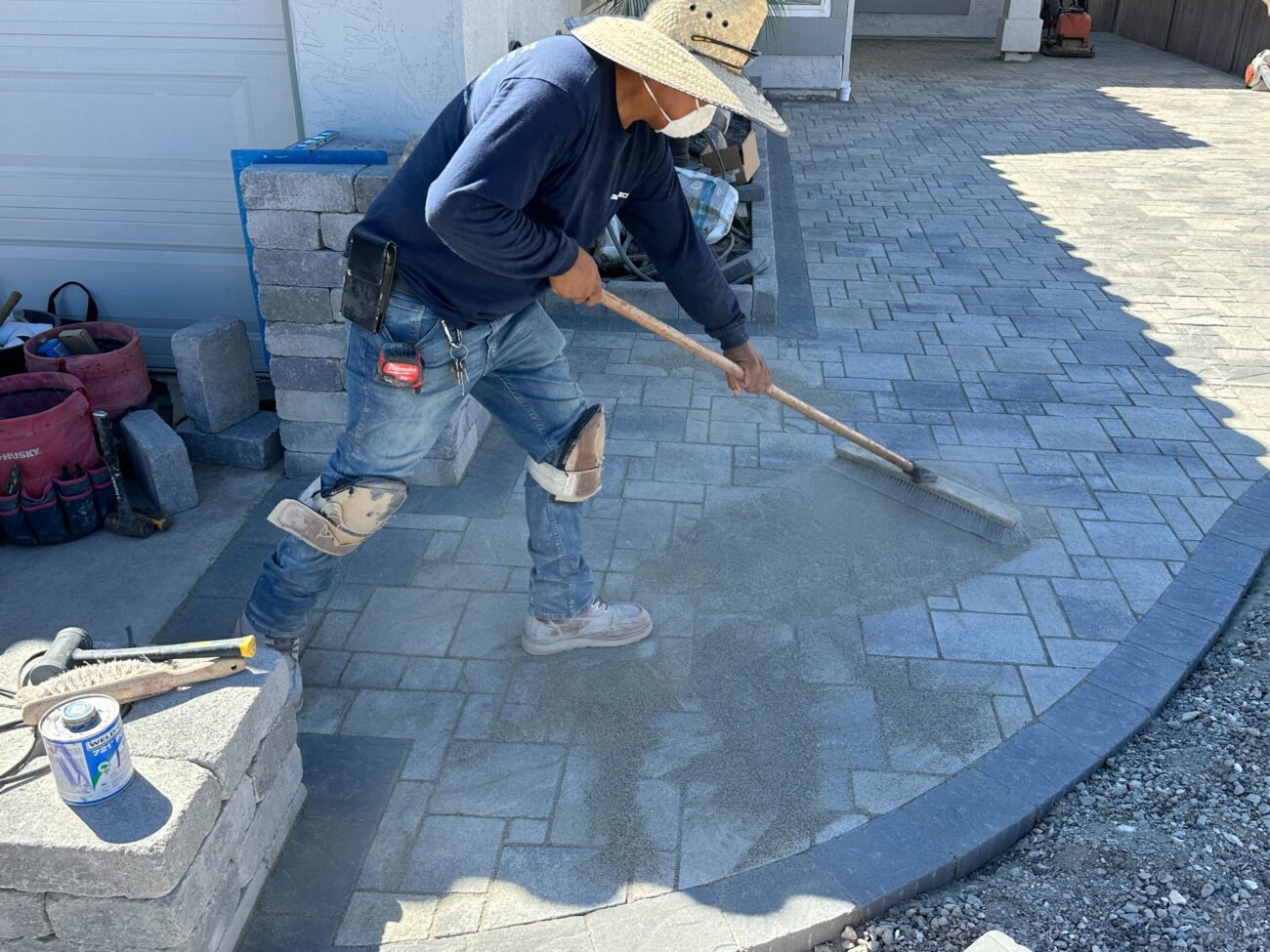 A crew member is sweeping jointing sand over newly installed interlocking pavers along a curved driveway/patio edge....