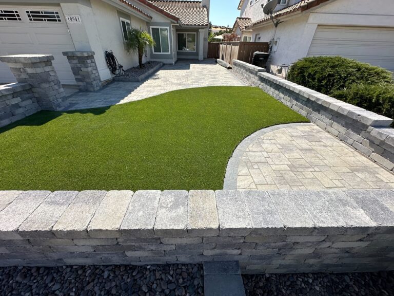 Residential front yard showing a finished interlocking paver patio and adjacent artificial turf. Segmental retaining...