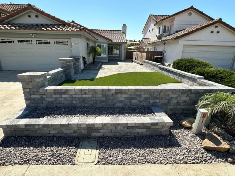 Front-yard site photo showing a completed segmental retaining wall with capstones forming a raised bed containing...