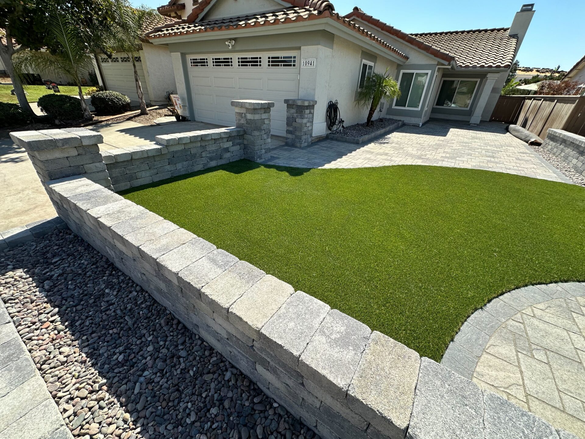 Front-yard residential hardscape showing completed interlocking paver driveway, walkway and patio with a curved segmental...