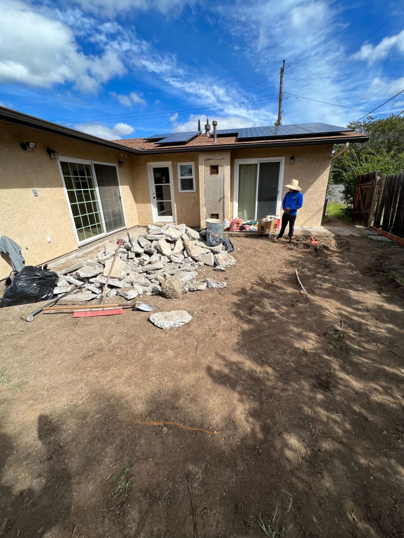 Residential backyard showing a demolished concrete patio with a large pile of broken concrete rubble and exposed soil. A...